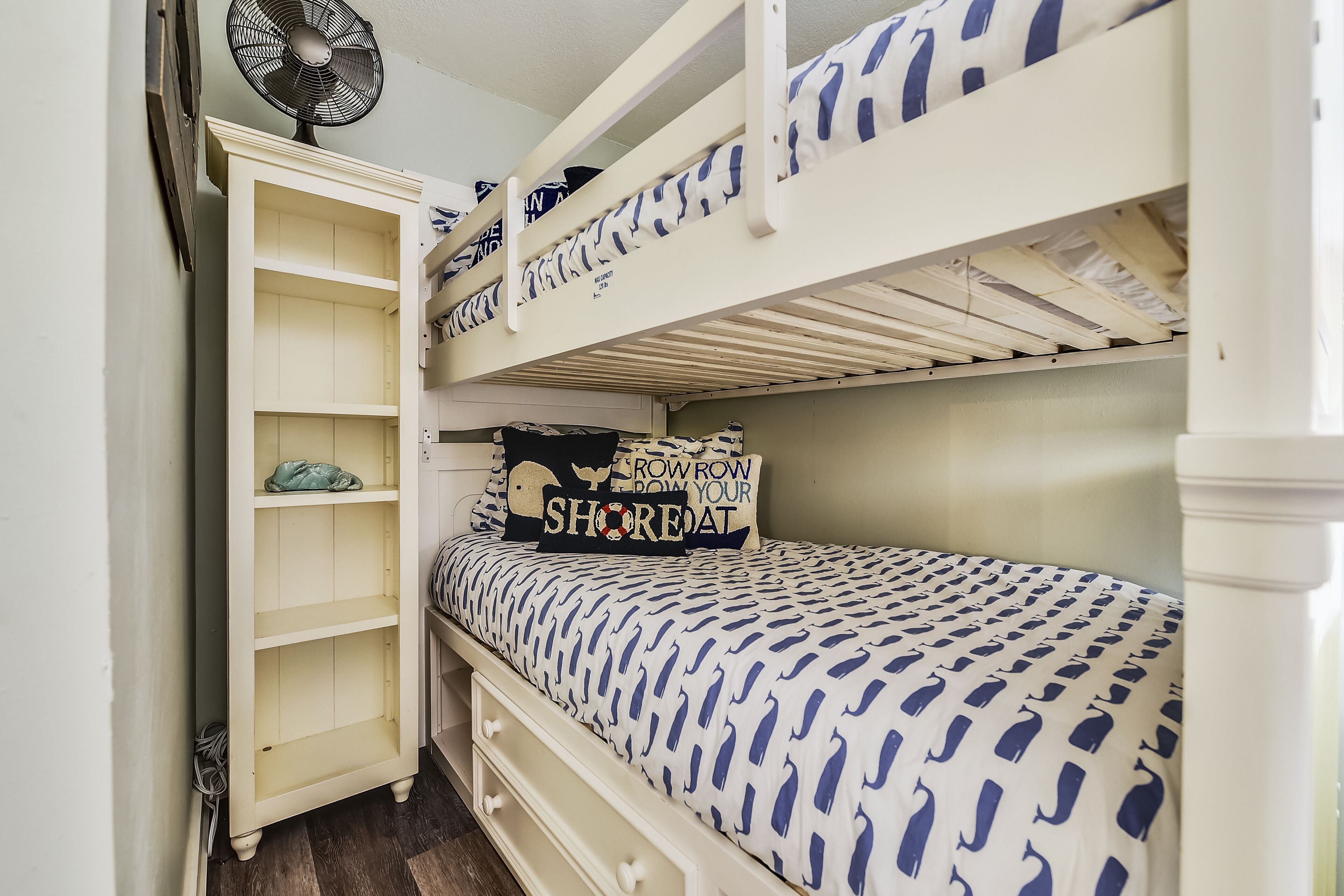 A bunk bed with a patterned bedspread, a shelf unit, and a fan.