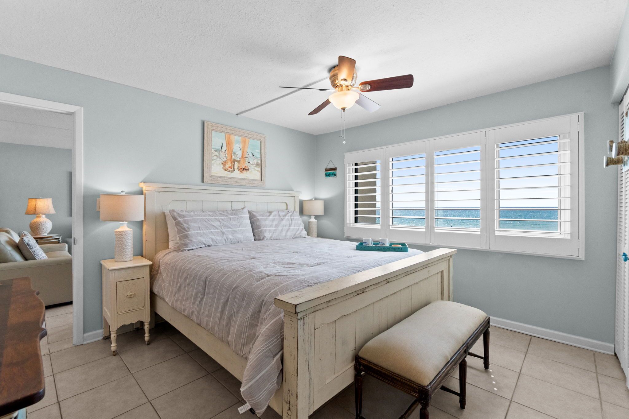 A bedroom with a bed, bedside tables, a ceiling fan, a window with shutters, and a view of the ocean.