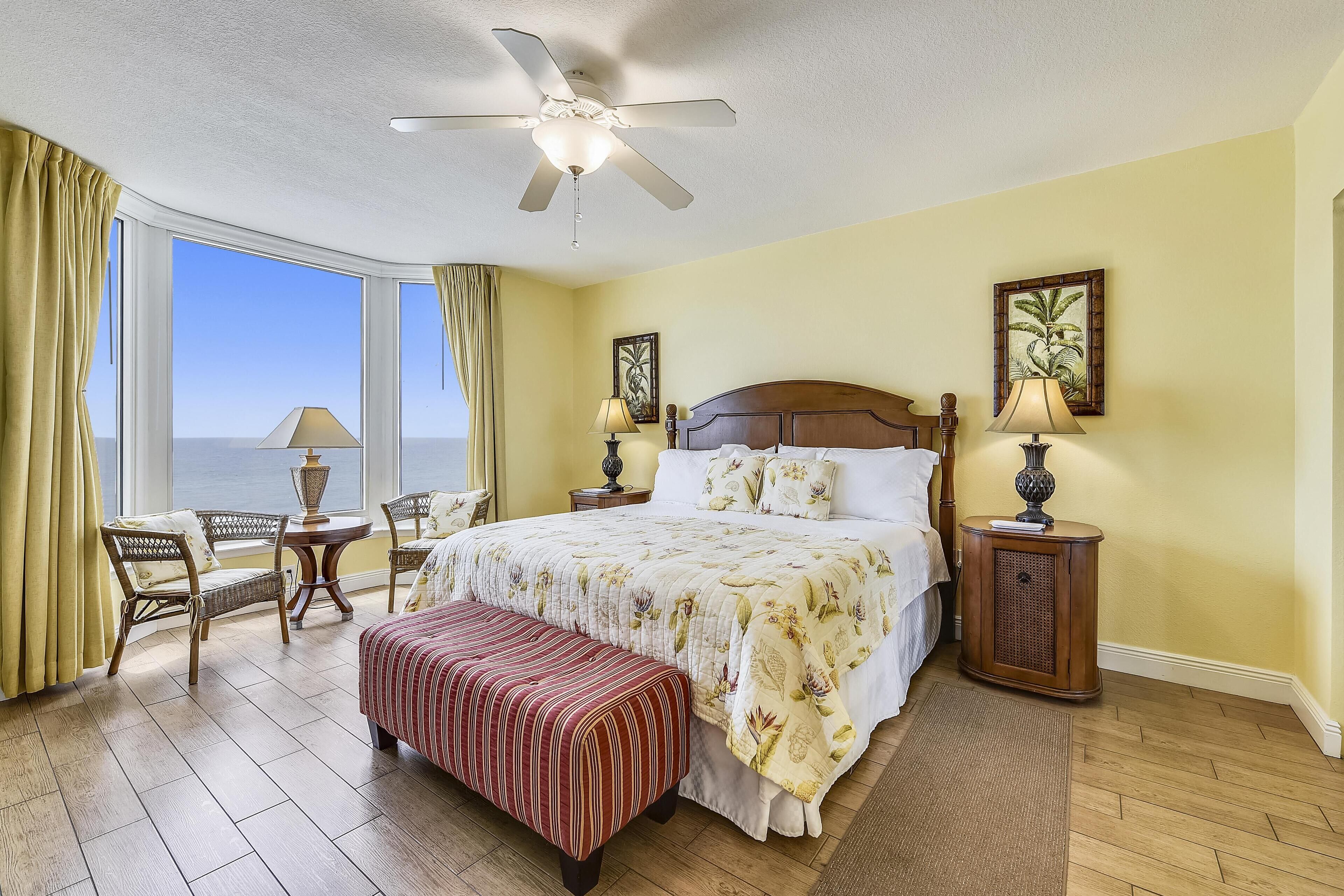 Une chambre d’hôtel avec un grand lit, un ventilateur de plafond, une vue sur l’océan, une fenêtre avec des rideaux et une petite table avec une lampe.