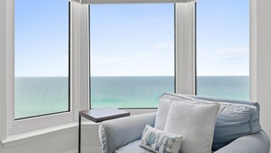 A room with a view of the ocean through large windows, a light blue sofa with a pillow, and a small table. - Emerald Beach by Book That Condo (Panama City Beach)