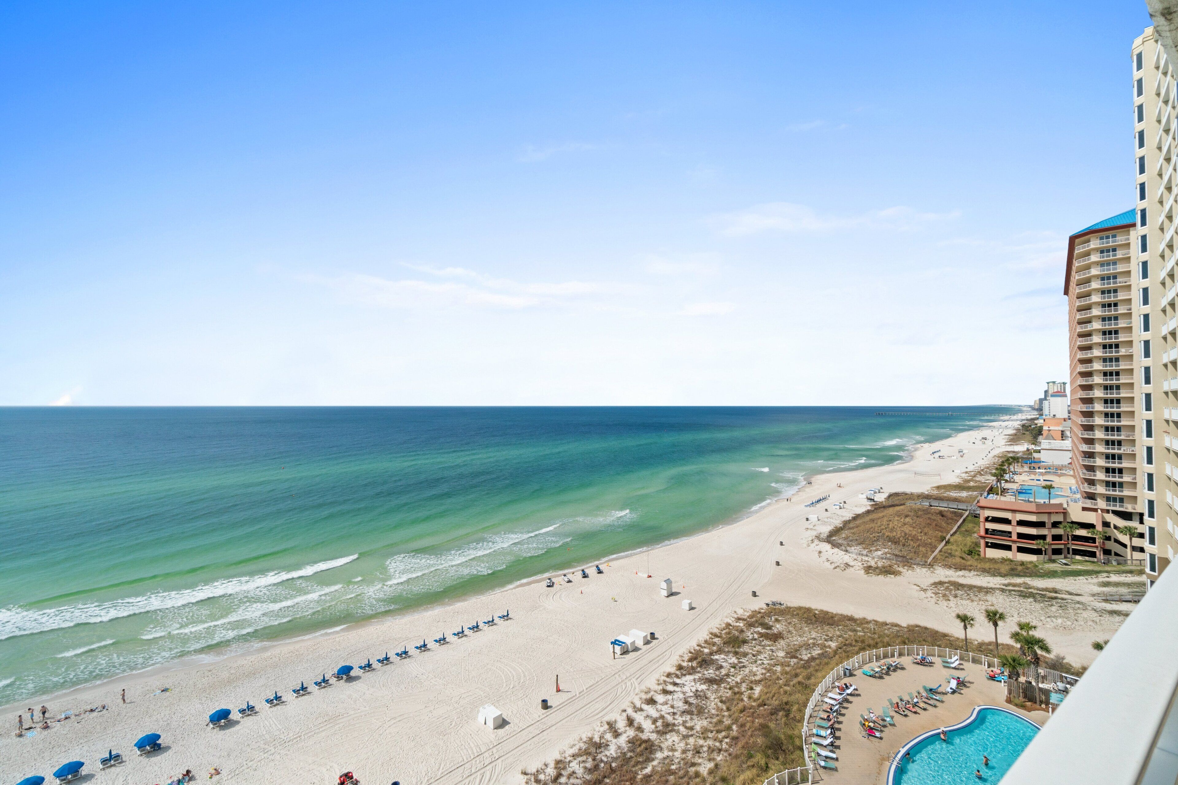 A beach view with a pool, beach umbrellas, and a tall building.