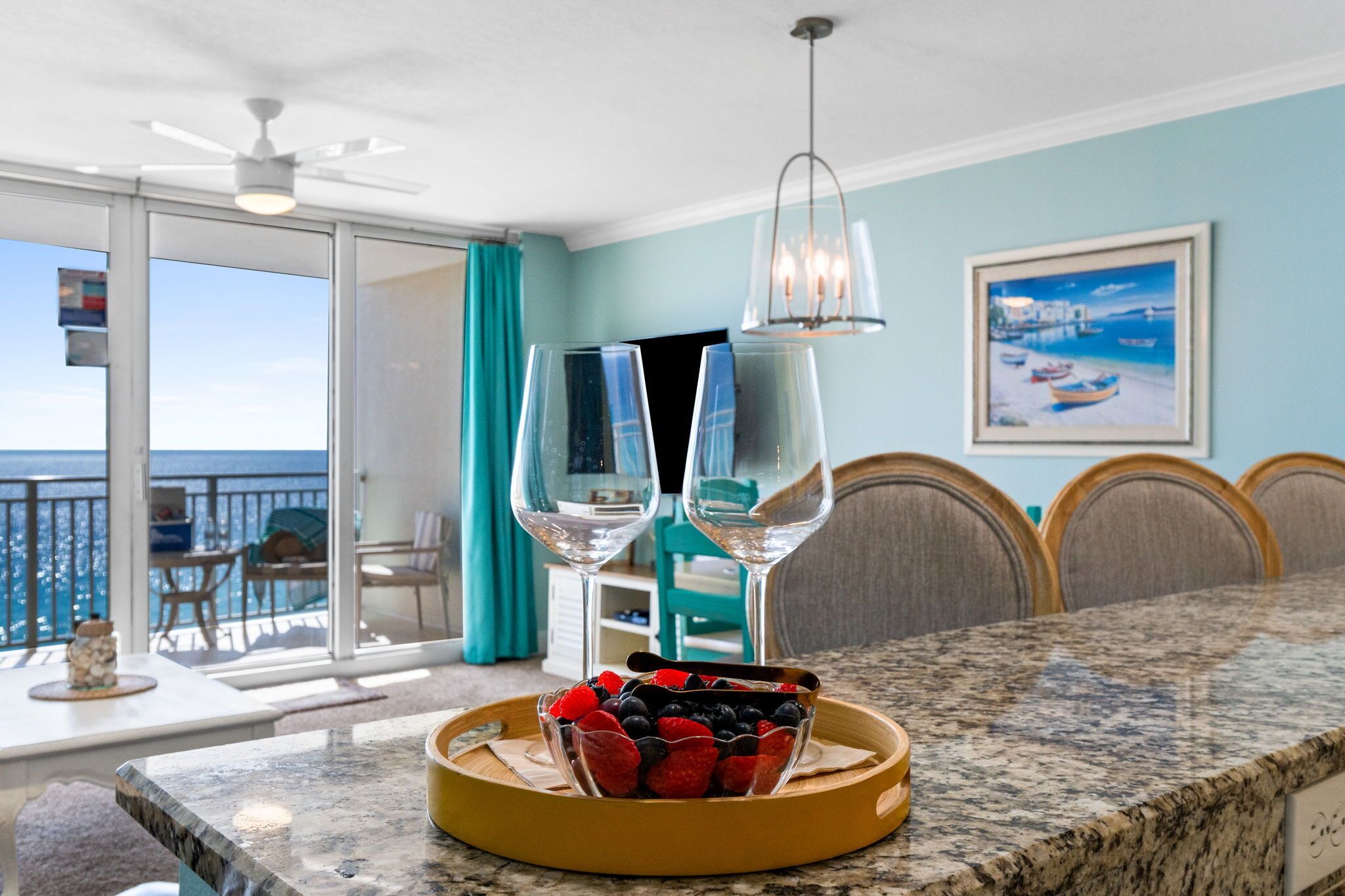 Uma área de refeições com mesa de buffet, duas taças de vinho, uma tigela de frutas e vista para o oceano por uma porta de correr de vidro.