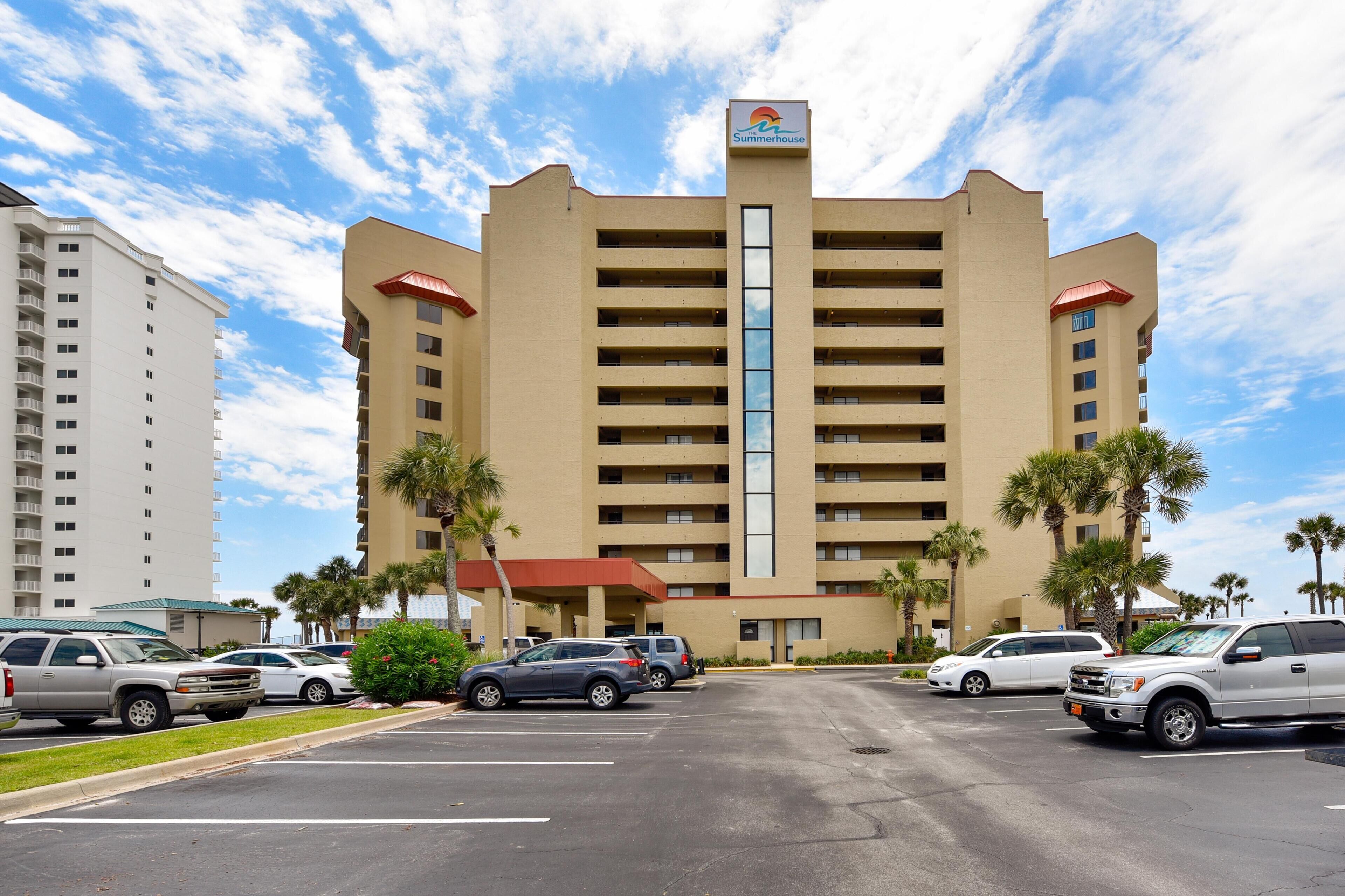 A hotel with a central vertical glass tower, multiple balconies, and a parking lot with several cars.