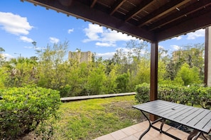 A covered patio with a table and benches, surrounded by lush greenery and a view of buildings in the distance.