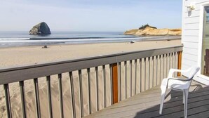 Una terraza de madera con una silla de plástico blanco, con vista a una playa arenosa y una formación rocosa con acantilados a lo lejos.