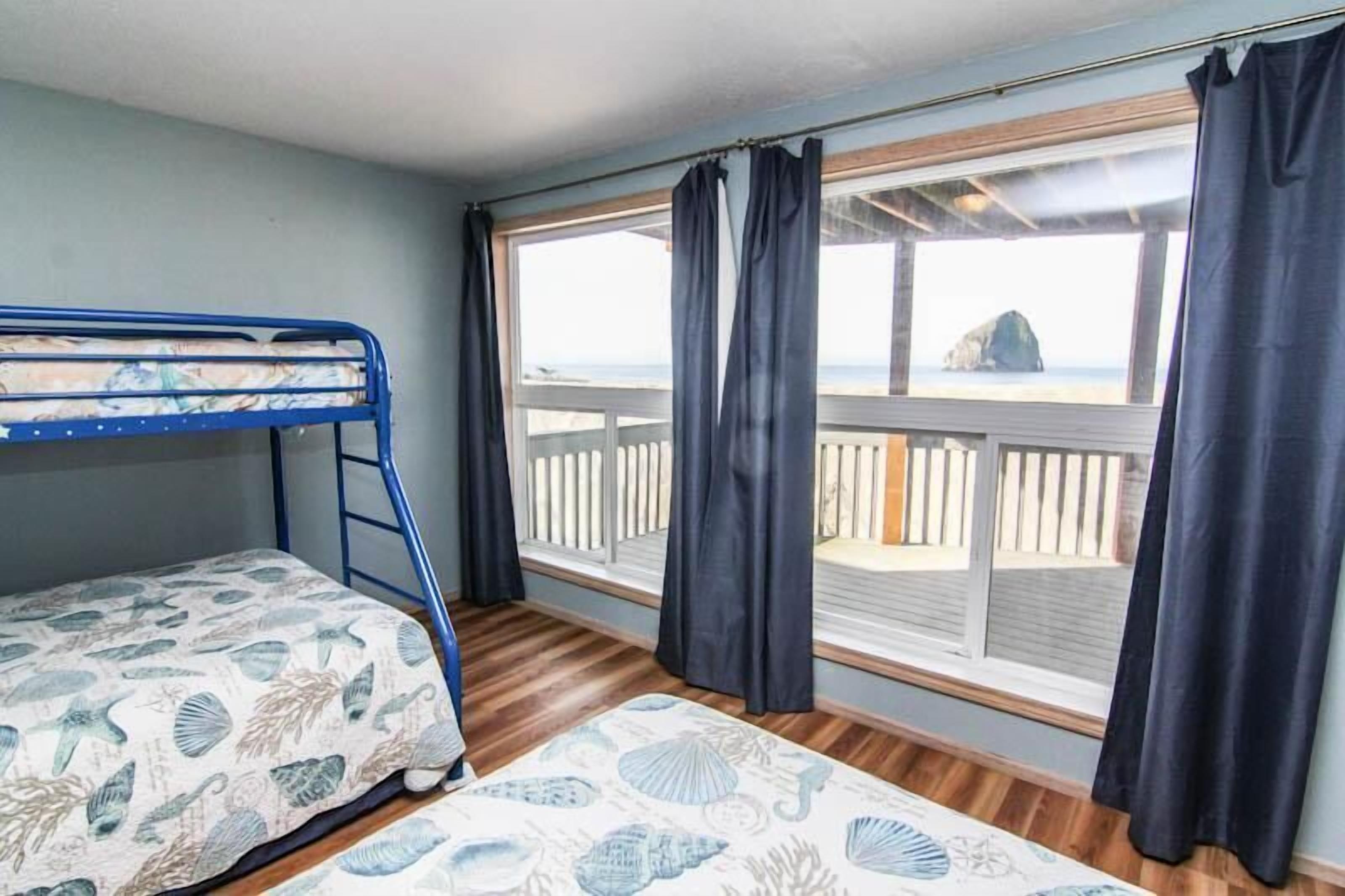 Une chambre avec un lit superposé, une grande fenêtre avec des rideaux, une terrasse en bois à l’extérieur et une vue sur la mer.