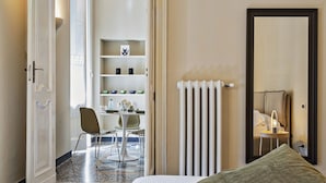A hotel room with a bed, a chair, a table with a vase of flowers, a radiator, and a shelf with decorative items. - Via Roma Luxury Apartment (Genova)