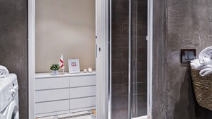 A modern bathroom with a glass shower enclosure, a white cabinet with toiletries, and a wicker basket with towels. - Via Roma Luxury Apartment (Genova)