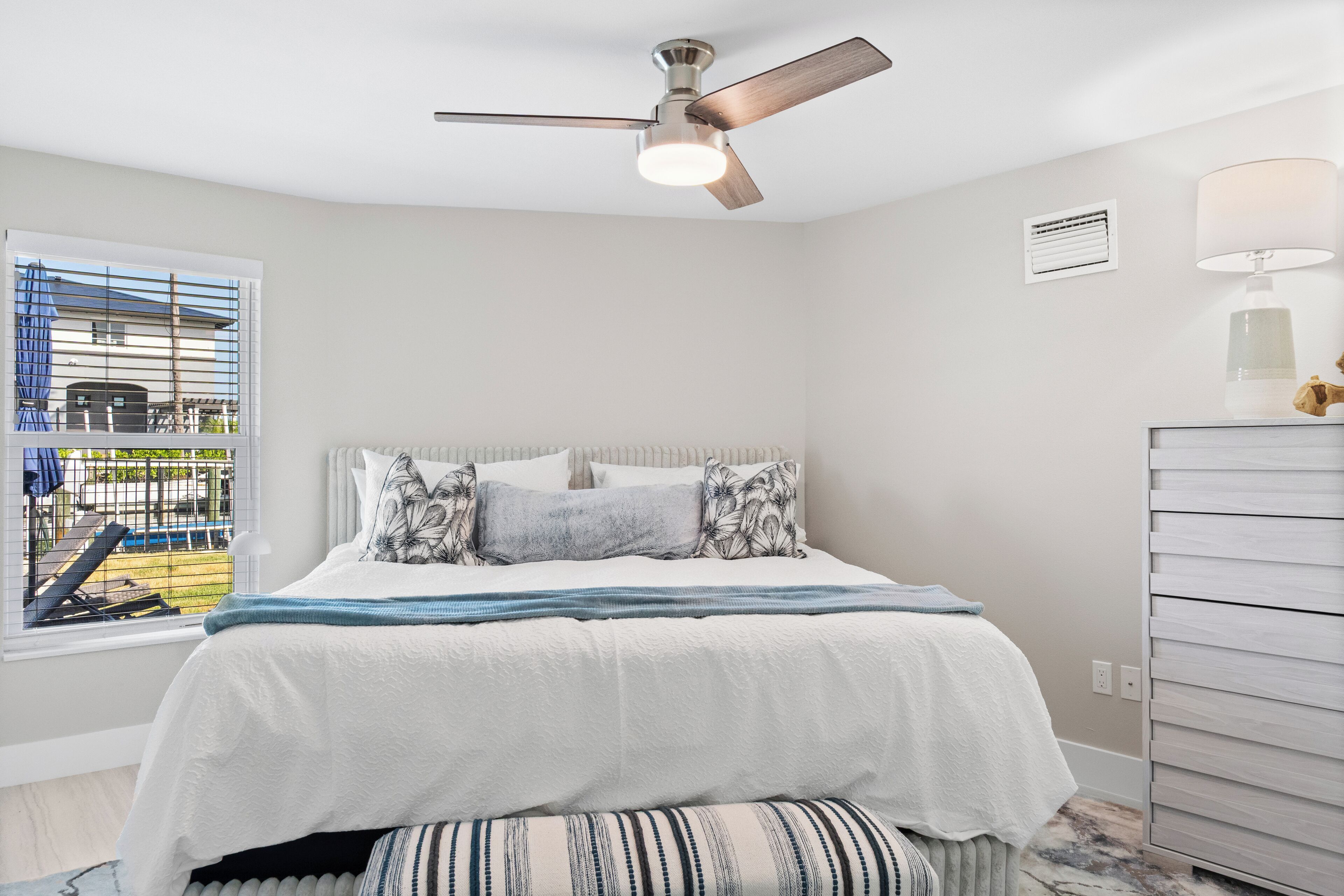 Un dormitorio con cama, ventilador de techo, ventana con persianas, lámpara y cómoda.