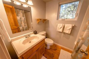A bathroom with a wooden vanity, a white sink, a toilet, and a window with a view of trees. - Cloud Nine (Depoe Bay)