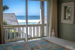 A bedroom with a large window overlooking the ocean, a wooden deck, and a bed with a floral bedspread. - Cloud Nine (Depoe Bay)