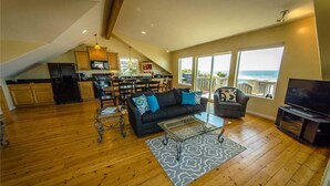 A spacious living area with a wooden floor, a black sofa with blue cushions, a glass coffee table, and a TV stand. - Cloud Nine (Depoe Bay)