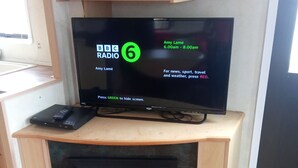 Un téléviseur diffusant la chaîne BBC Radio 6, une télécommande et un ordinateur portable sont posés sur une étagère en bois au-dessus d’une cheminée.