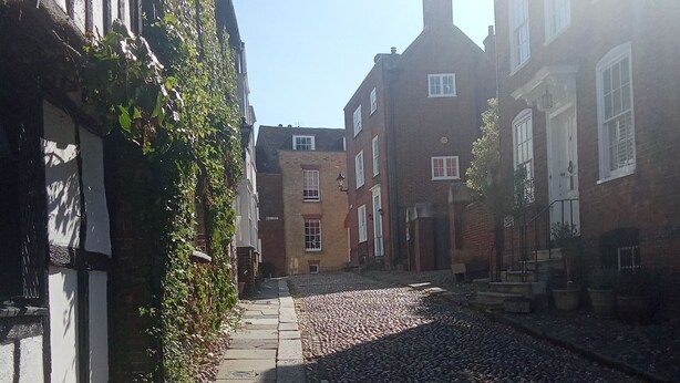 Une rue pavée bordée de bâtiments historiques, avec un mélange de façades en briques et de structures en bois, et un ciel dégagé au-dessus.