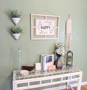 A wall with a framed sign saying "THIS IS OUR HAPPY PLACE", two potted plants, a macrame wall hanging, a glass table with a white basket, a vase with flowers, a framed photo, and a small decorative item.