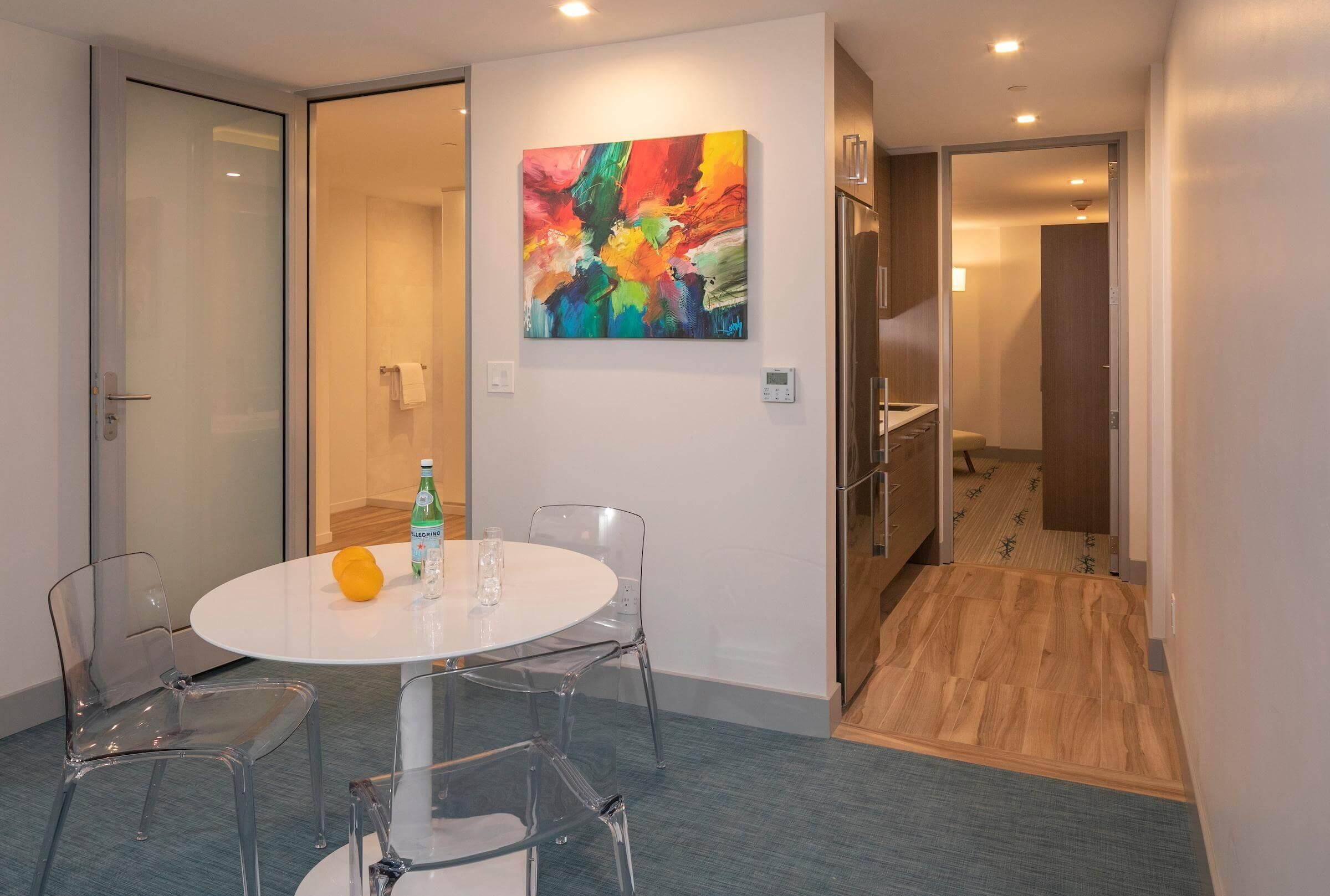 Une chambre d’hôtel moderne avec une table ronde, des chaises transparentes, un tableau abstrait aux couleurs vives et un plancher en bois.