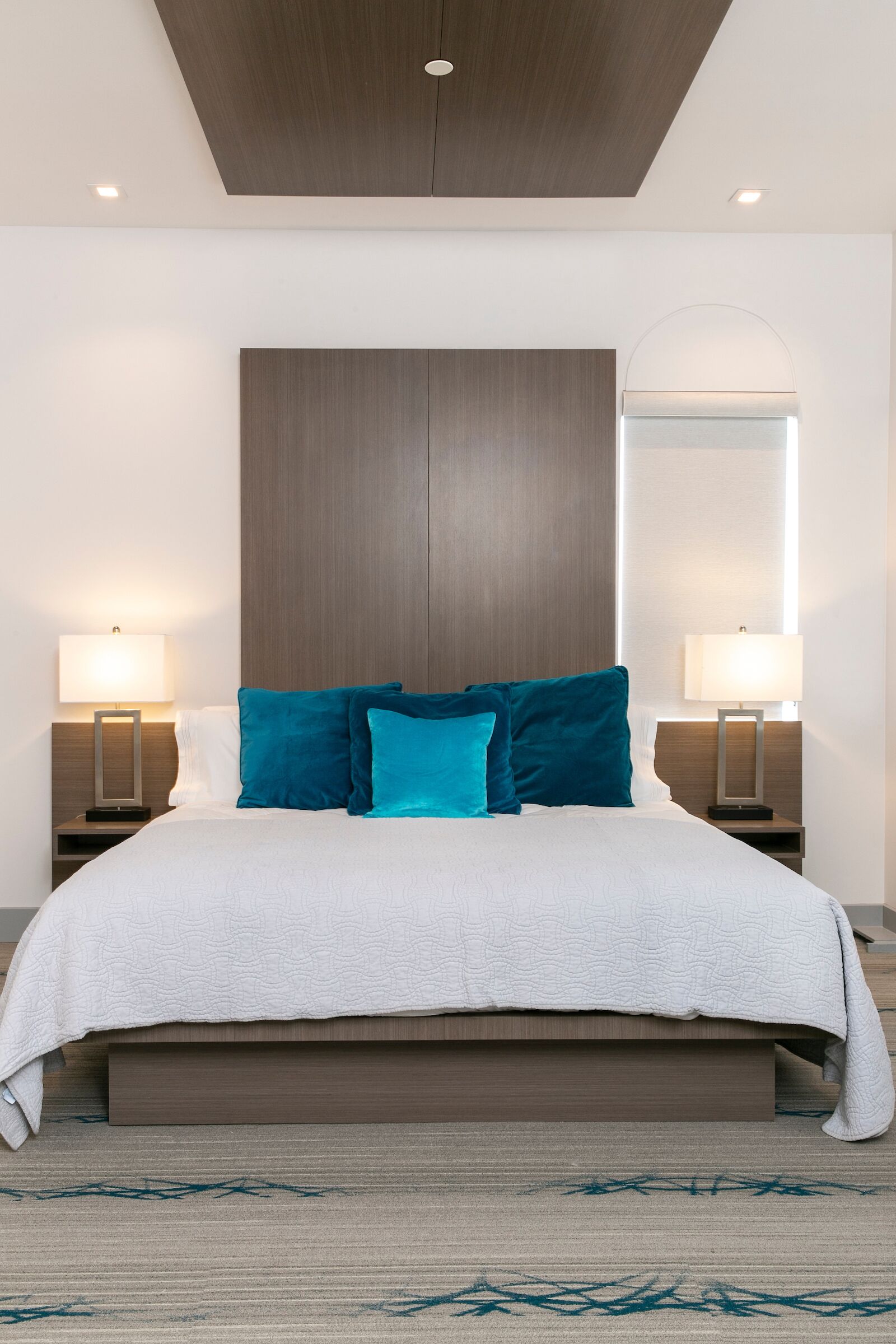 A modern hotel room with a large bed, two bedside lamps, a wooden headboard, and a patterned carpet.