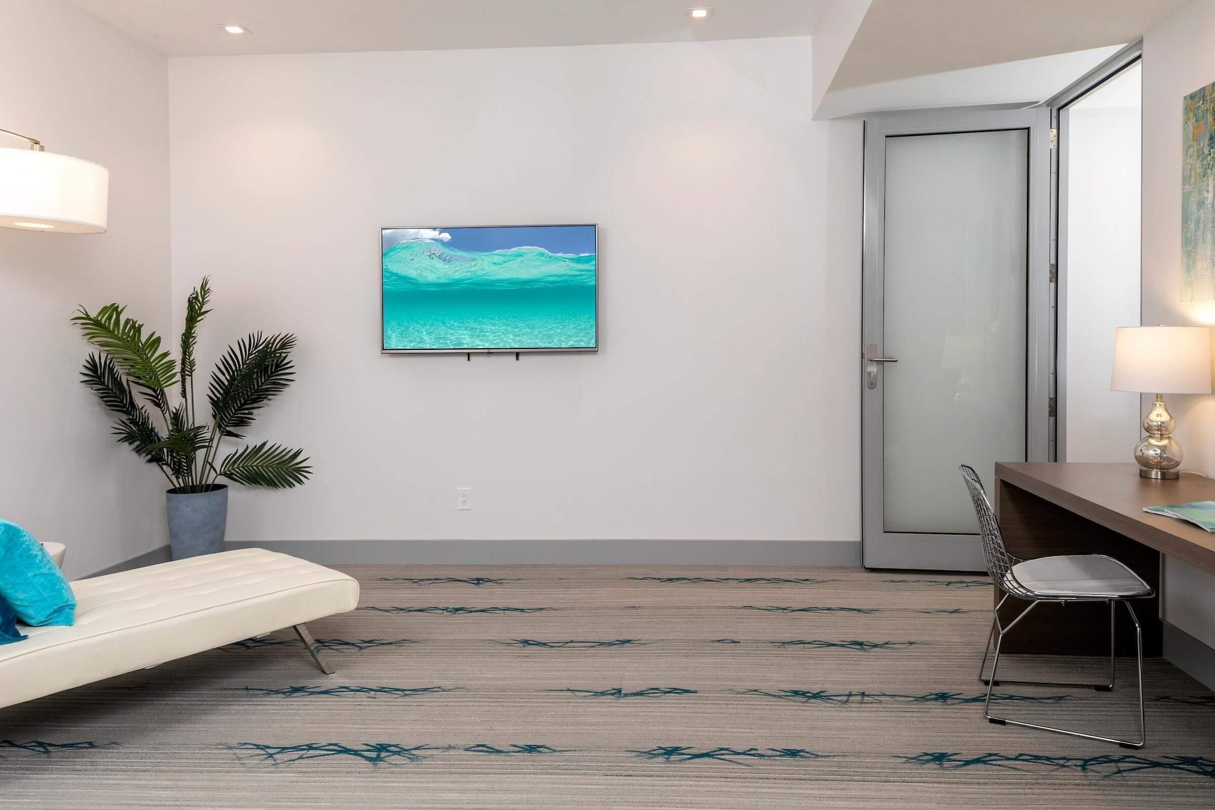 A modern hotel room with a minimalist design, featuring a bed, a desk, a chair, a potted plant, and a large window with a view of the ocean.