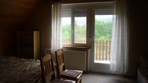 A bedroom with a wooden ceiling, a bed, two chairs, a table, and a window with a view of greenery.