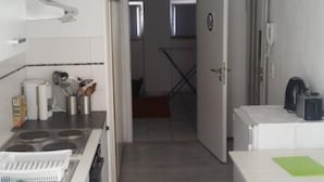 A narrow kitchen with a stove, sink, and cabinets. There is a small table with a green placemat and a kettle on the counter. A door leads to a hallway with a radiator and a window. - Fewo Mayer am Bodensee (Friedrichshafen)