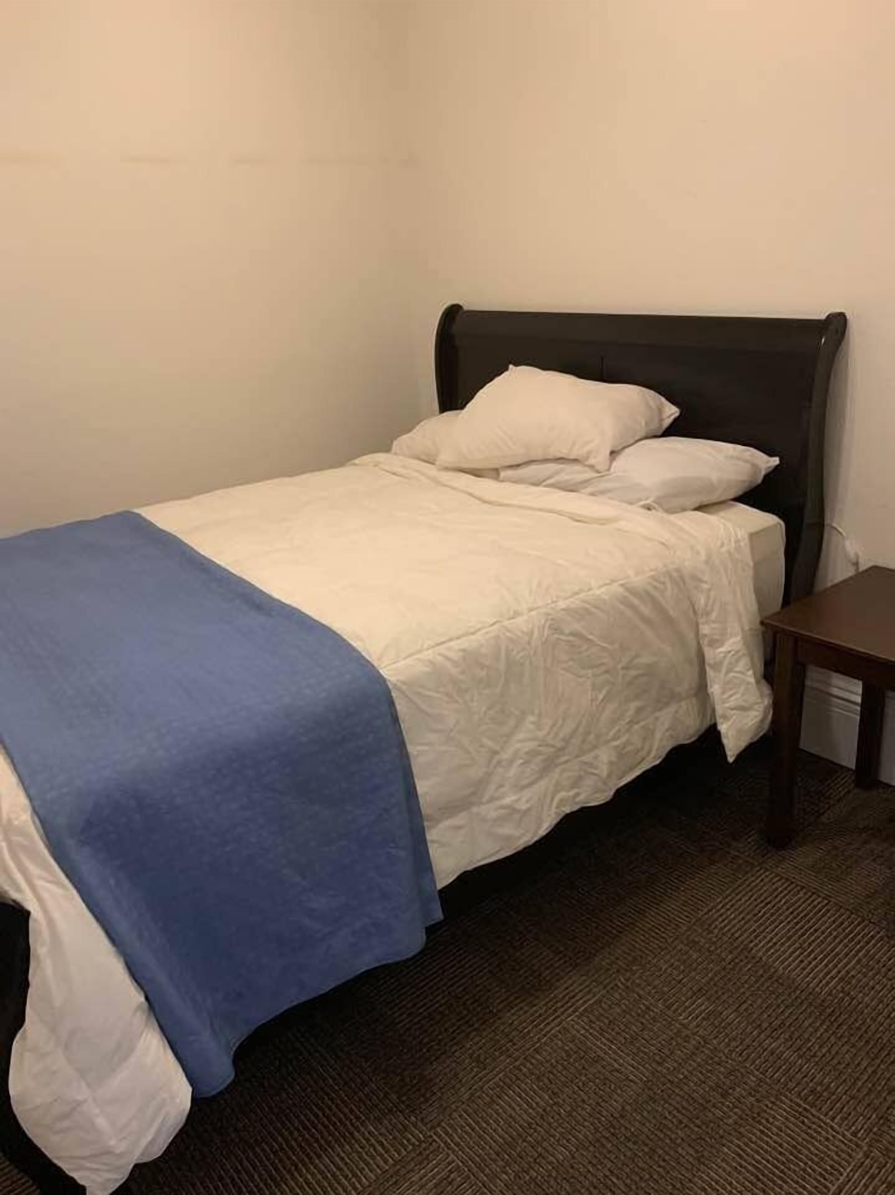 Una cama individual con cabecera oscura, ropa de cama blanca y una manta azul.