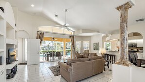 A spacious living room with a large beige sectional sofa, a ceiling fan, and a view of the outdoor patio area. - Set Sail for a Spectacular Adventure - Gulf Access, Pool, Kayaks - Villa Sentio - Roelens Vacations (Cape Coral)