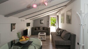 Een moderne woonkamer met een grijze bank, een glazen eettafel met een schaal fruit, en een plafond met zichtbare balken en roze bolvormige lampen.