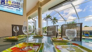 Une table dressée pour le dîner, avec de l’argenterie colorée et des verres à vin, une enseigne indiquant « Roelens Vacations », et une vue sur des palmiers et une piscine.