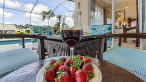 Une table avec une assiette de fraises et un verre de vin rouge, donnant sur une piscine et des palmiers.