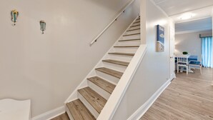 Un escalier avec des marches en bois et des murs blancs, menant à une pièce où se trouvent une chaise blanche et une table.