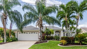 Una casa de dos pisos con garaje, rodeada de un césped cuidado y diversas plantas tropicales.