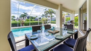 Mesa de vidrio con sillas de mimbre, con vista a una piscina y a palmeras y casas.