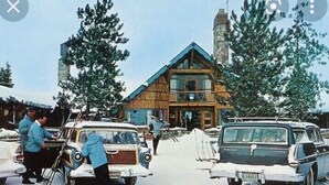 Una cabaña de madera con balcón, rodeada de nieve y coches estacionados.