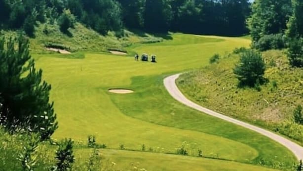 Un campo de golf con un sendero serpenteante, rodeado de un bosque frondoso y bajo un cielo parcialmente nublado.