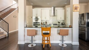 Cocina con armarios blancos, campana extractora de acero inoxidable y refrigerador de acero inoxidable. Hay dos taburetes de bar y una silla de madera.