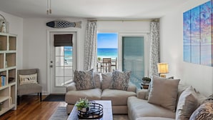 A living room with a sofa, a coffee table with decorative items, a bookshelf, and a view of the ocean through a door. - BEACHFRONT Beach Vacation! Eat Breakfast While You Watch Dolphins!! (Destin)