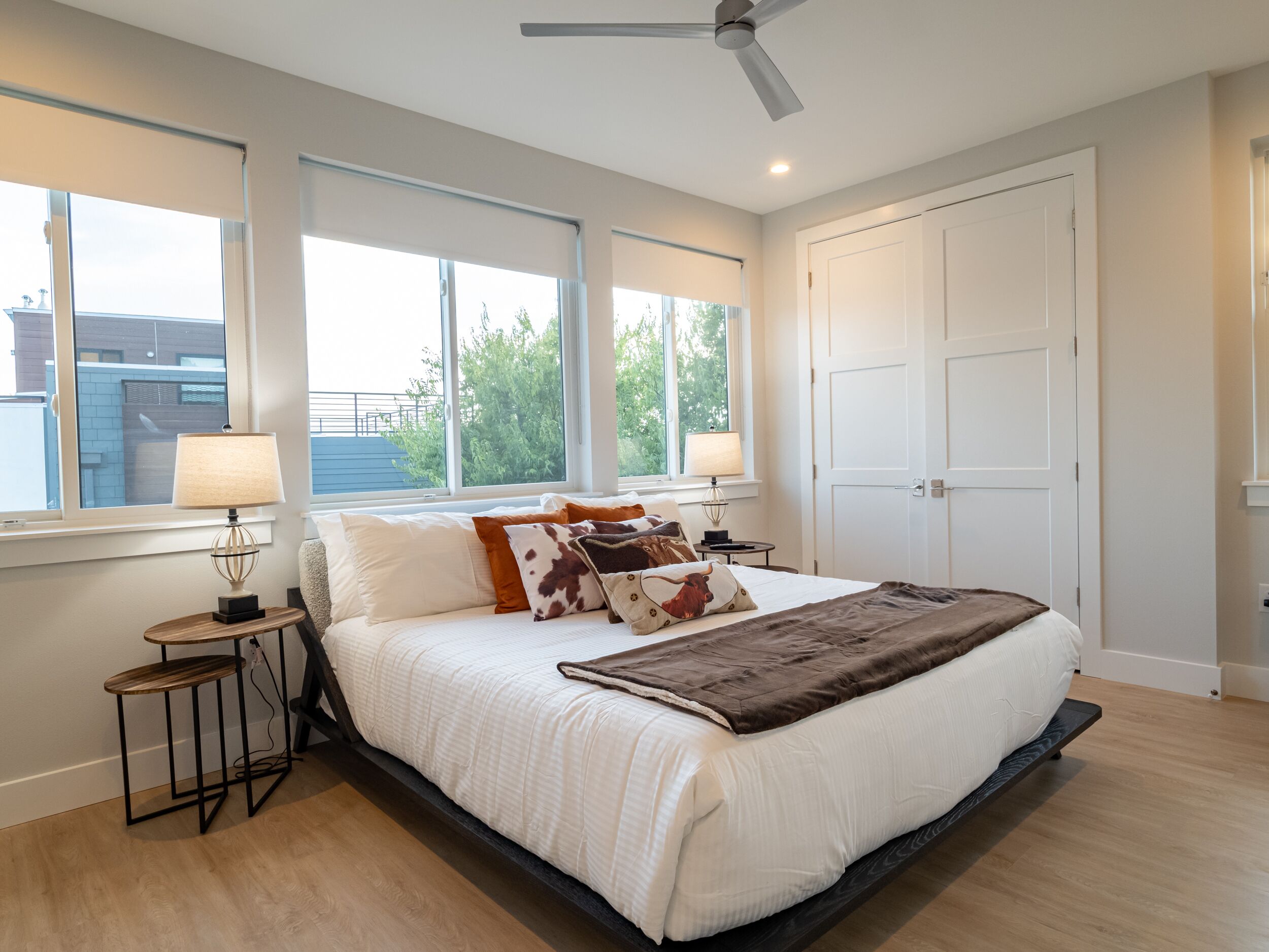 A bedroom with a large bed, two bedside tables, a ceiling fan, and a view of the outside.