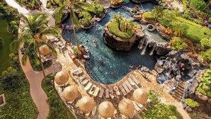 Een resort zwembad met verschillende waterattracties, waaronder watervallen en een centraal zwembadgedeelte omringd door ligbedden en parasols.
