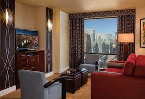 A hotel room with a large window, a flat-screen TV, a red sofa, a grey armchair, and a small table with a lamp.