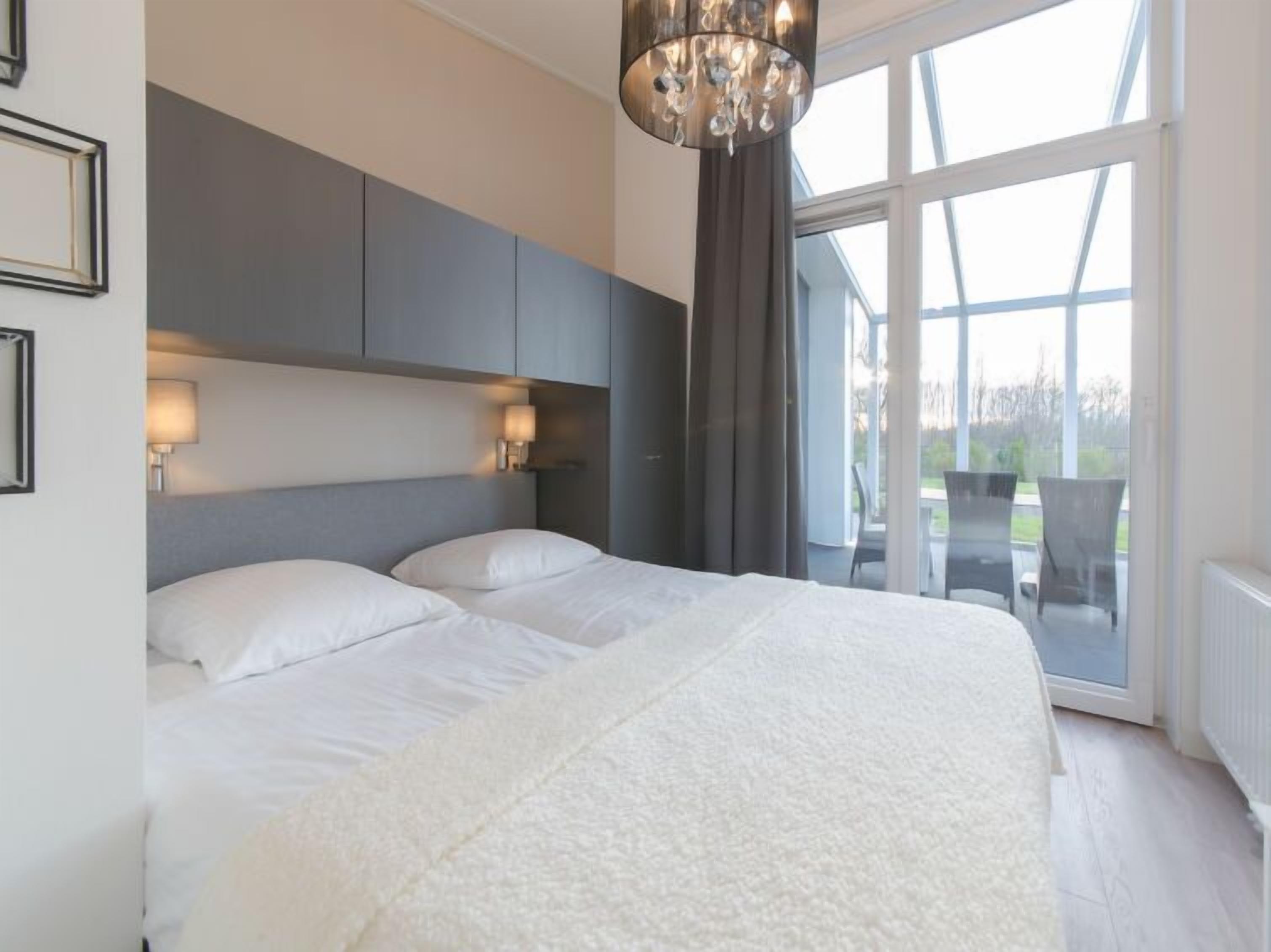 Ein modernes Schlafzimmer mit einem großen Bett, einer Kronleuchter und Blick ins Freie durch große Fenster.