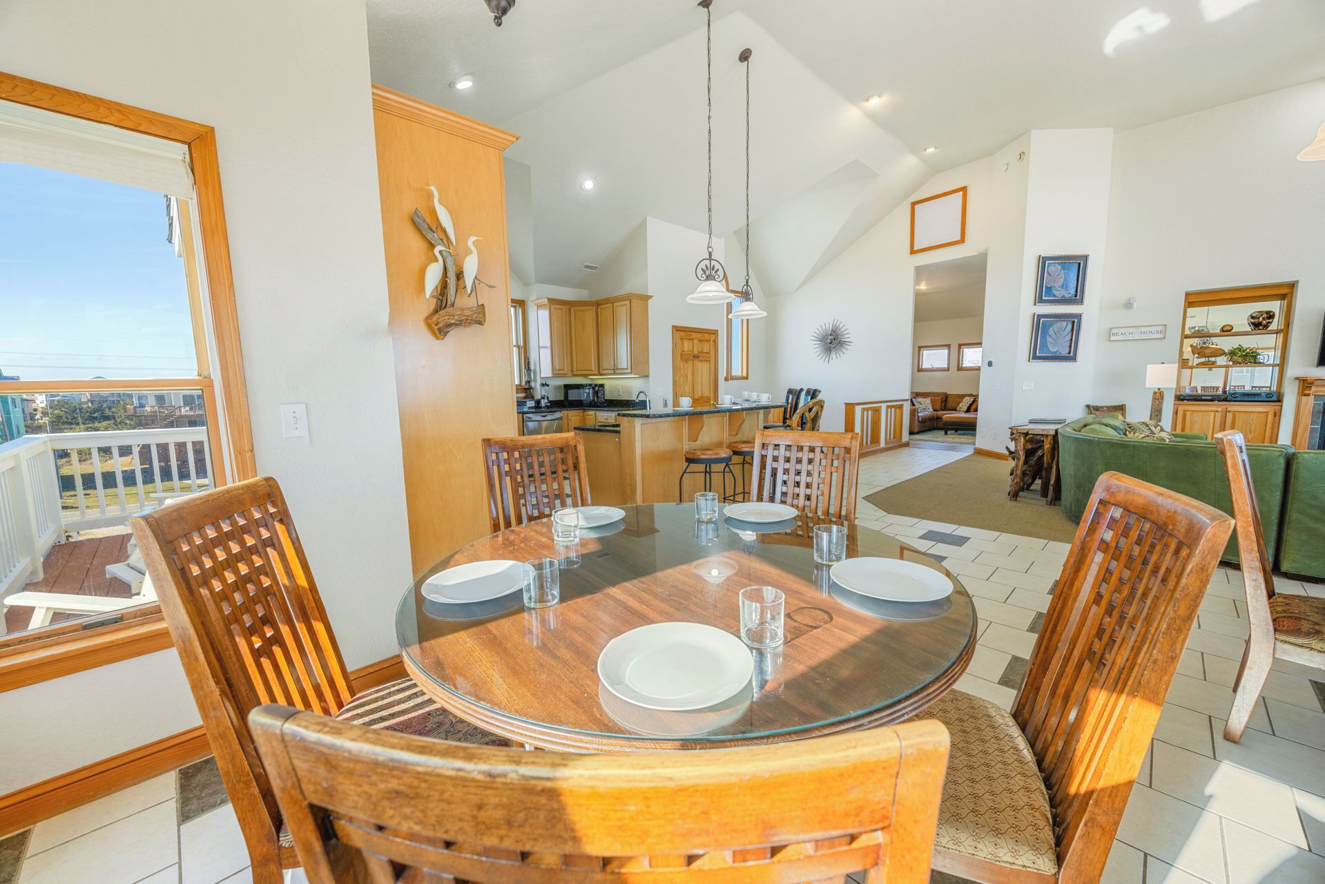 A spacious dining area with a round glass table, wooden chairs, and a view of the kitchen and living room in the background.