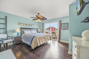 Une chambre avec un lit, deux tables de chevet, une chaise, un ventilateur de plafond et une vue sur une maison rouge, visible à travers une porte vitrée.