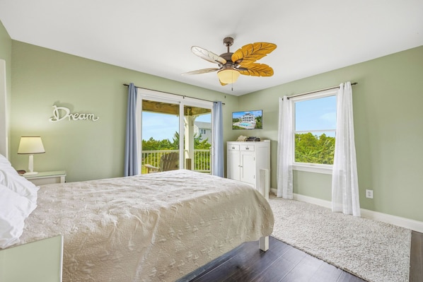 A bedroom with a bed, a ceiling fan, a nightstand, a dresser, a window with curtains, and a balcony with a chair.