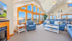 A spacious living room with a fireplace, large windows, and a ceiling fan. - Oceanview w/ Private Pool, Hot Tub, Game Room, and Wet Bar. Surf's Up Rodanthe! (Rodanthe)