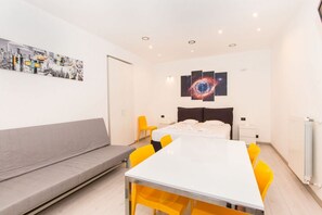 A modern hotel room with a bed, a dining table with chairs, a sofa, and a wall with a cityscape and a galaxy-themed artwork.