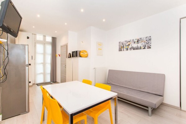 A modern kitchen with a white table and yellow chairs, a grey sofa, and a flat-screen TV mounted on the wall.
