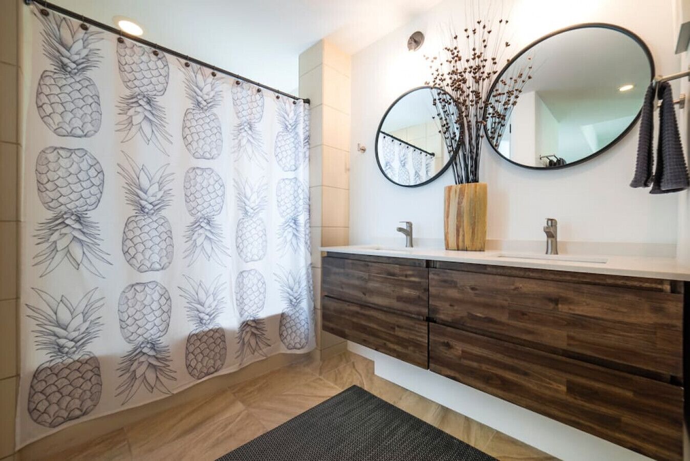 Une salle de bain avec un rideau de douche orné de motifs ananas, un meuble-lavabo en bois avec un miroir et un petit vase contenant des fleurs séchées.