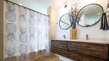 Une salle de bain avec un rideau de douche orné de motifs ananas, un meuble-lavabo en bois avec un miroir et un petit vase contenant des fleurs séchées.