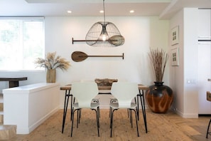A dining area with a wooden table, white chairs, a hanging light fixture, and a vase with tall plants. - ContemporaryHawaiianReno in Wailea*Pool*Beach*Golf (Wailea)