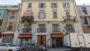 A multi-story building with a ground-level shop, a balcony with hanging plants, and a parked car.